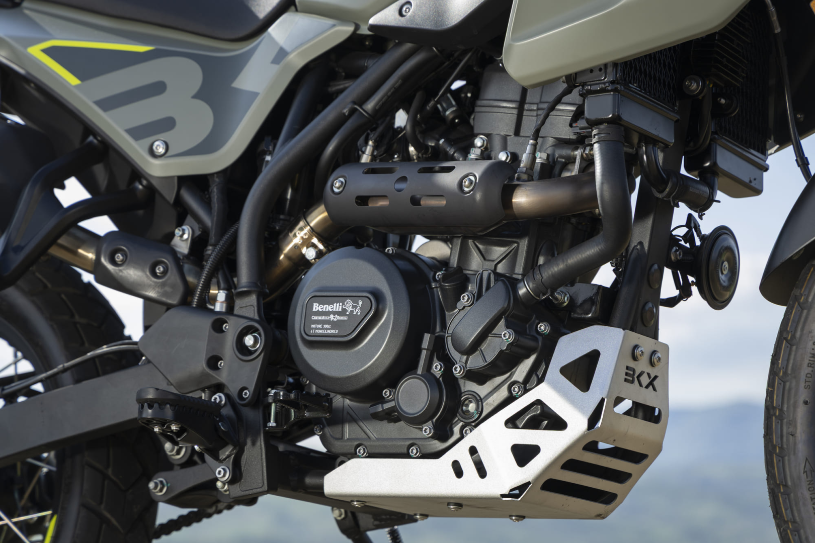 Benelli-bkx-300 2 - AutoBike Close-up of a Benelli motorcycle engine and frame, showing the exhaust pipe, heat shield, and skid plate nearby in a rugged setup. - AutoBike