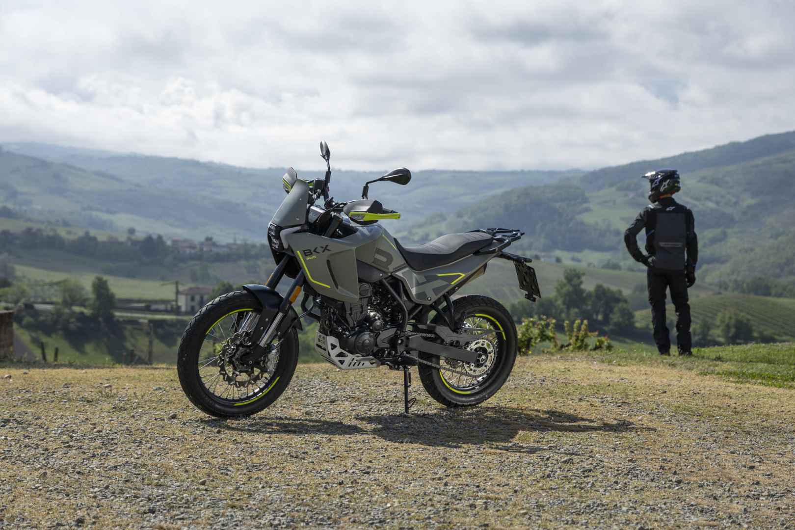 Benelli-bkx-300 3 - AutoBike Adventure motorcycle with gray and lime accents parked on a gravel area; a rider in black gear stands nearby, looking at rolling green hills in the distance. - AutoBike
