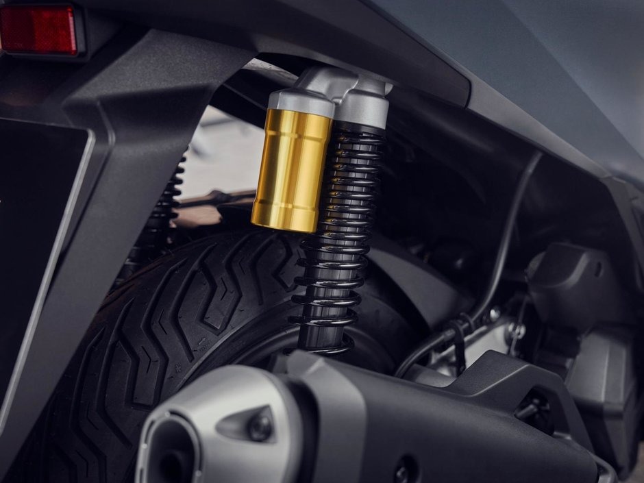 honda-pcx-125- 19 - AutoBike Gold-colored shock absorber and black coil spring of a motorcycle rear suspension beside a tire and exhaust area. - AutoBike