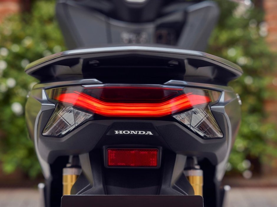 honda-pcx-125- 20 - AutoBike Rear of a gray Honda motorcycle with a glowing red tail light and visible Honda emblem on the back. - AutoBike