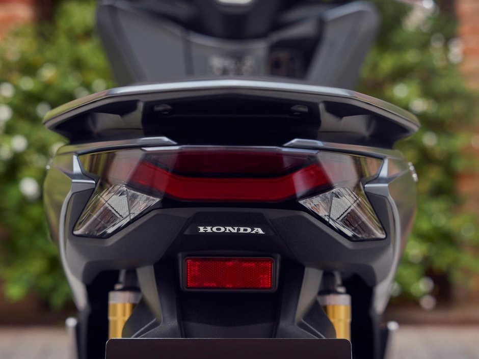 honda-pcx-125- 22 - AutoBike Rear view of a Honda motorcycle with distinctive twin taillights, red central reflector, and gold suspension visible beneath the tail. - AutoBike