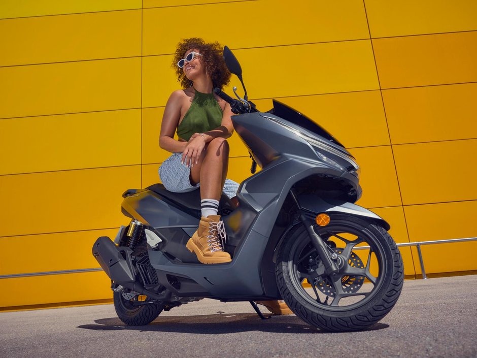 honda-pcx-125- 25 - AutoBike Woman sits on a gray scooter beside a vivid yellow wall, wearing a green halter top, denim shorts, and tan boots with striped socks. - AutoBike