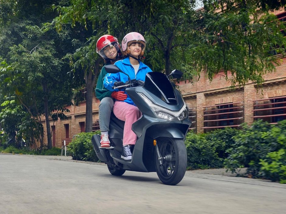 honda-pcx-125- 26 - AutoBike Two people wearing helmets ride a dark gray scooter along a tree-lined street. - AutoBike