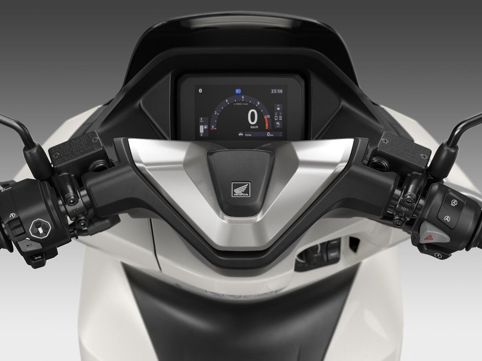 honda-pcx-125- 27 - AutoBike Rider’s view of a motorcycle dashboard: digital speedometer shows 0 km/h, Honda emblem in center, left and right handlebars with controls visible - AutoBike