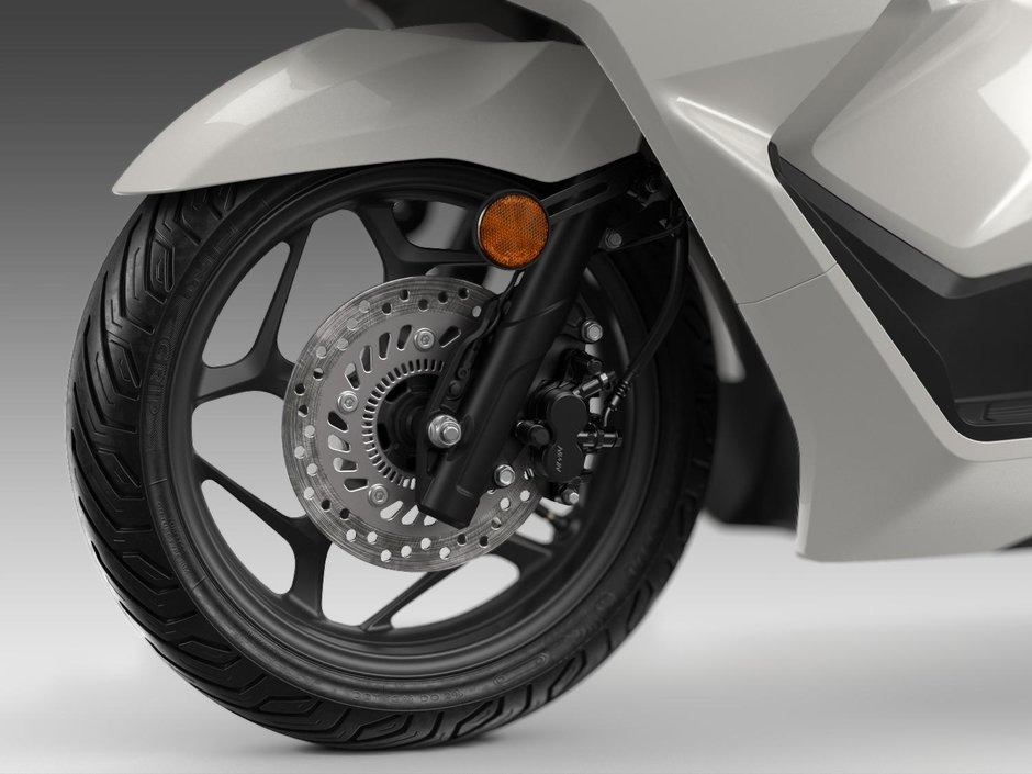honda-pcx-125- 29 - AutoBike Front wheel of a white motorcycle showing the disc brake, caliper, and orange reflector on the fork. - AutoBike