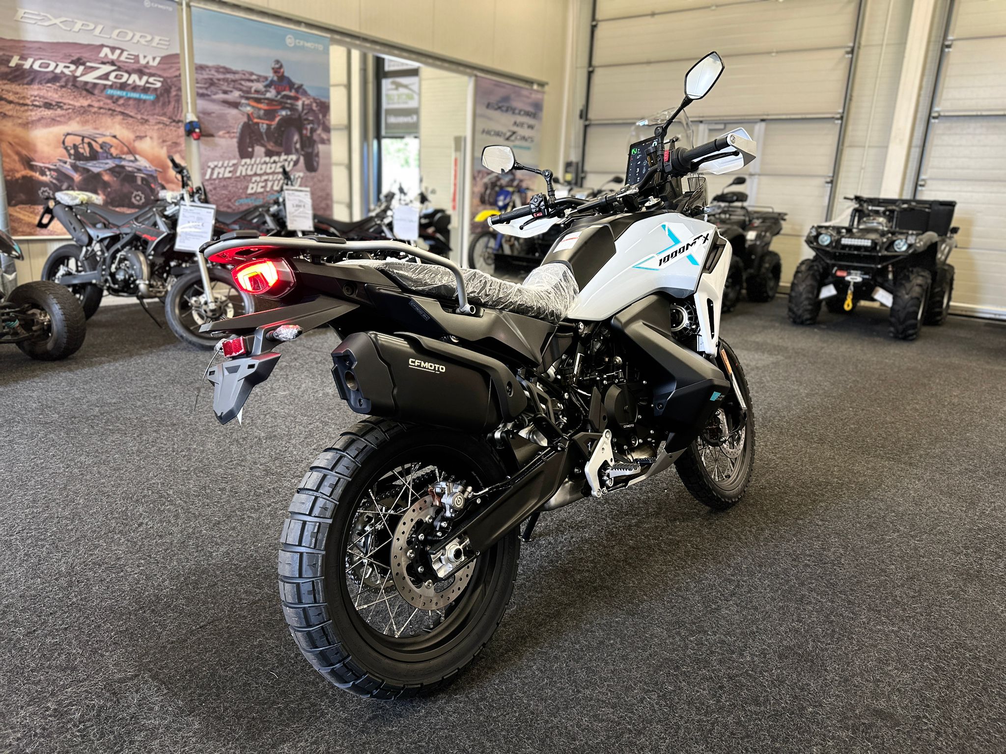 CFMOTO-1000-MT-X-1-18 at 203522 1 - AutoBike White CFmoto 1000MTX adventure bike in a showroom, with other motorcycles and promotional posters in the background. - AutoBike