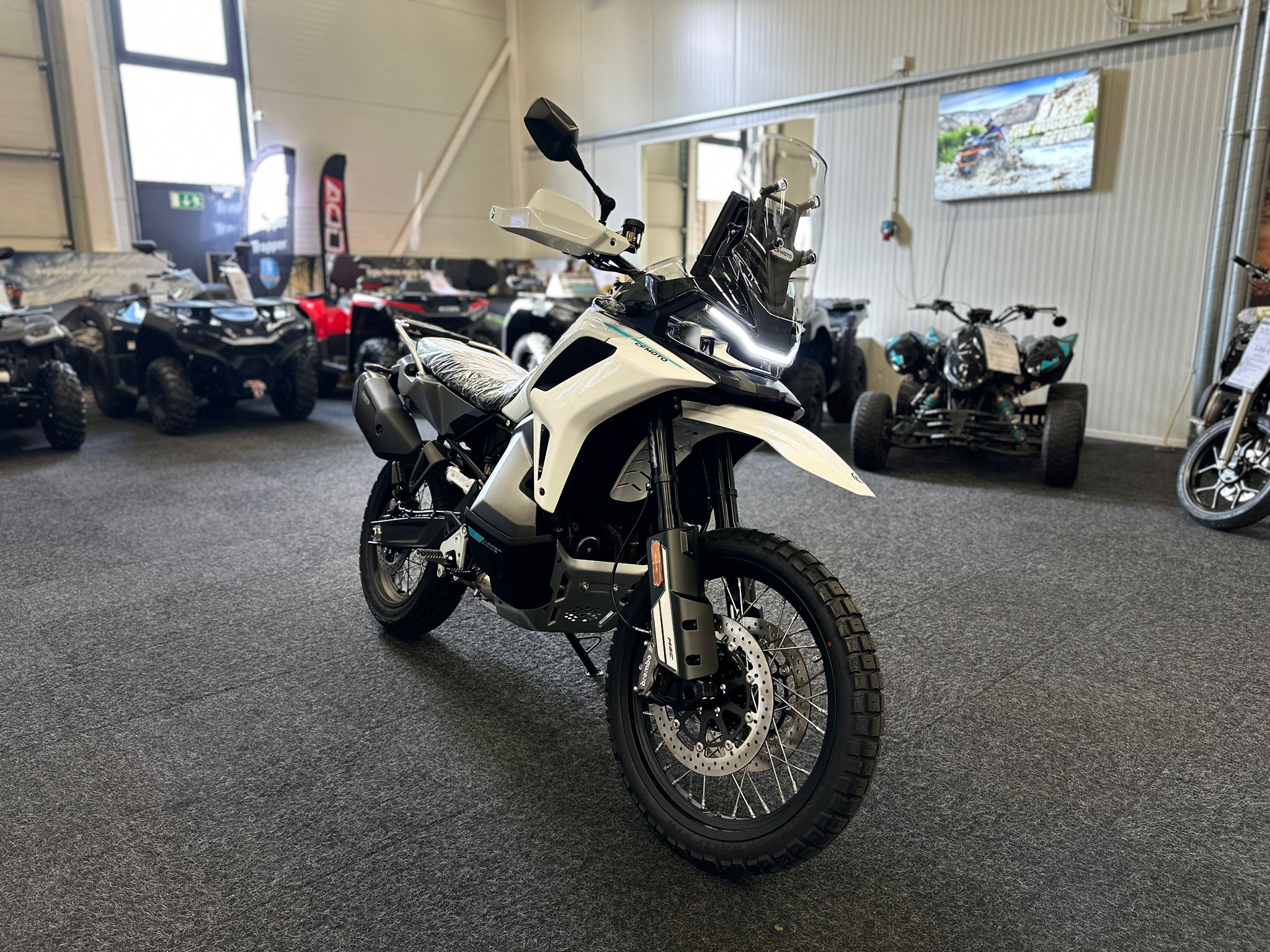 CFMOTO-1000-MT-X-1-18 at 203522 2 - AutoBike White adventure motorcycle in a showroom, with other ATVs and banners visible in the background - AutoBike