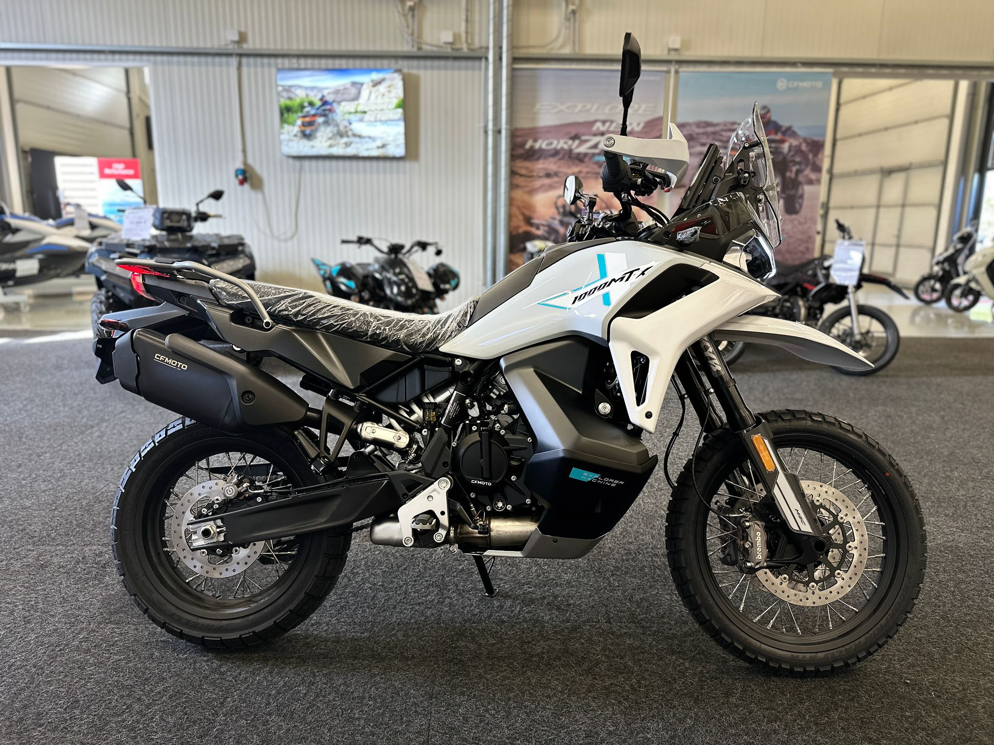 CFMOTO-1000-MT-X-1-18 at 203522 3 - AutoBike White and black adventure motorcycle on a showroom floor, with other bikes and posters in the background, rear wheel visible. - AutoBike