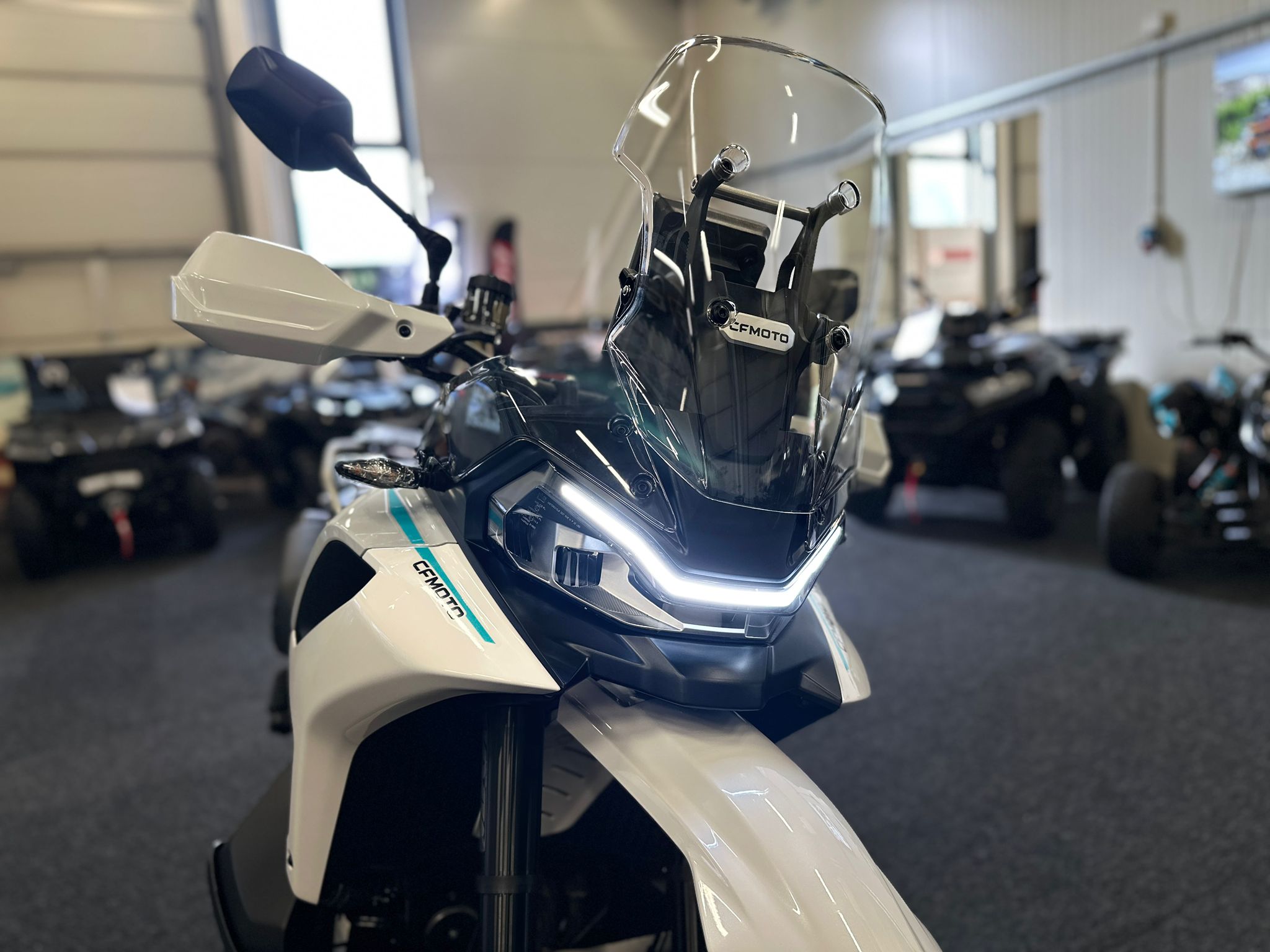 CFMOTO-1000-MT-X-1-18 at 203523 2 - AutoBike Close-up of a white adventure motorcycle front with a clear windscreen and LED strip, in a showroom setting. - AutoBike