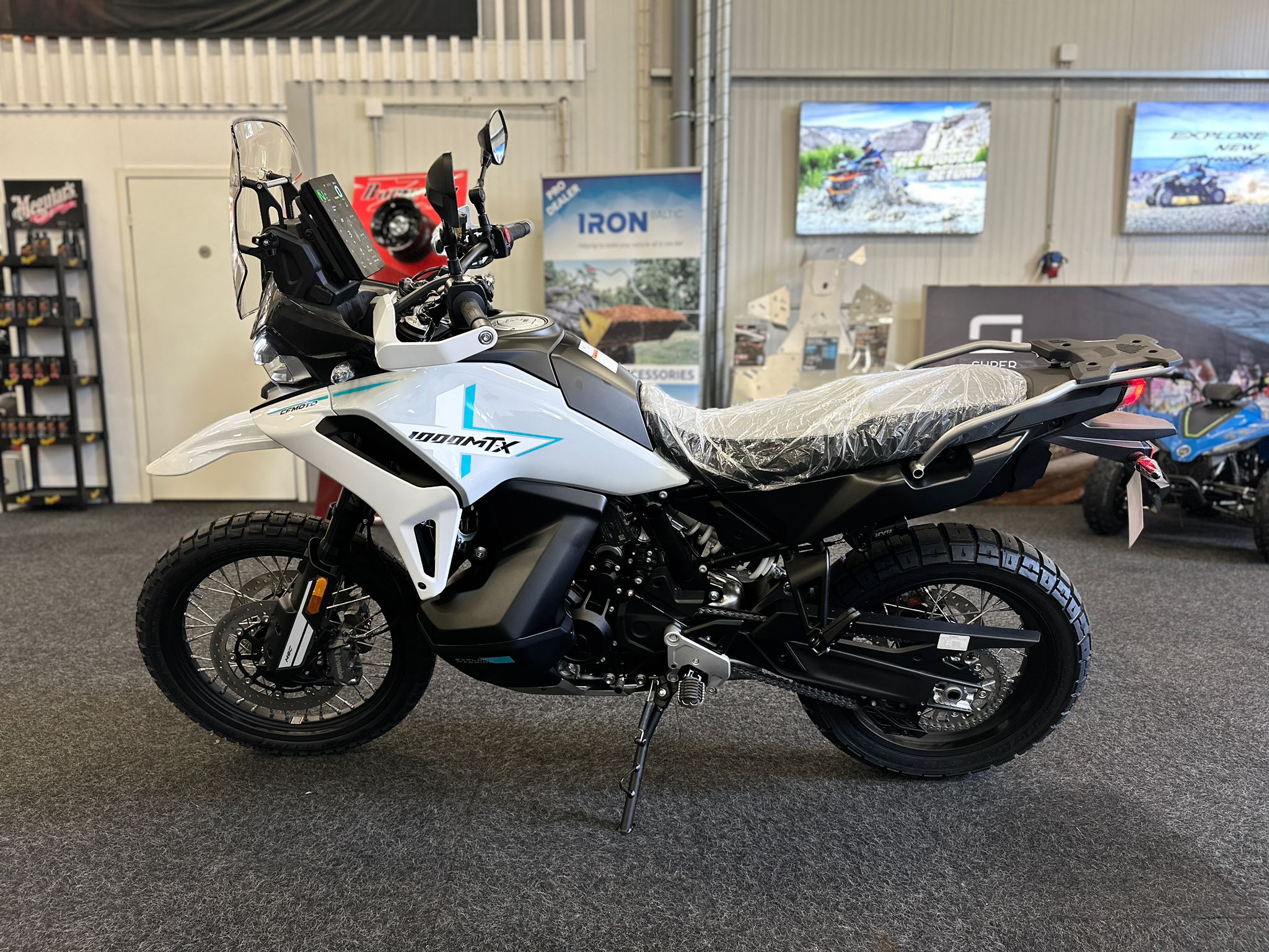 CFMOTO-1000-MT-X-1-18 at 203523 3 - AutoBike White and black adventure motorcycle on a display stand in a showroom, with a tall windshield and knobby tires, posters on the back wall. - AutoBike