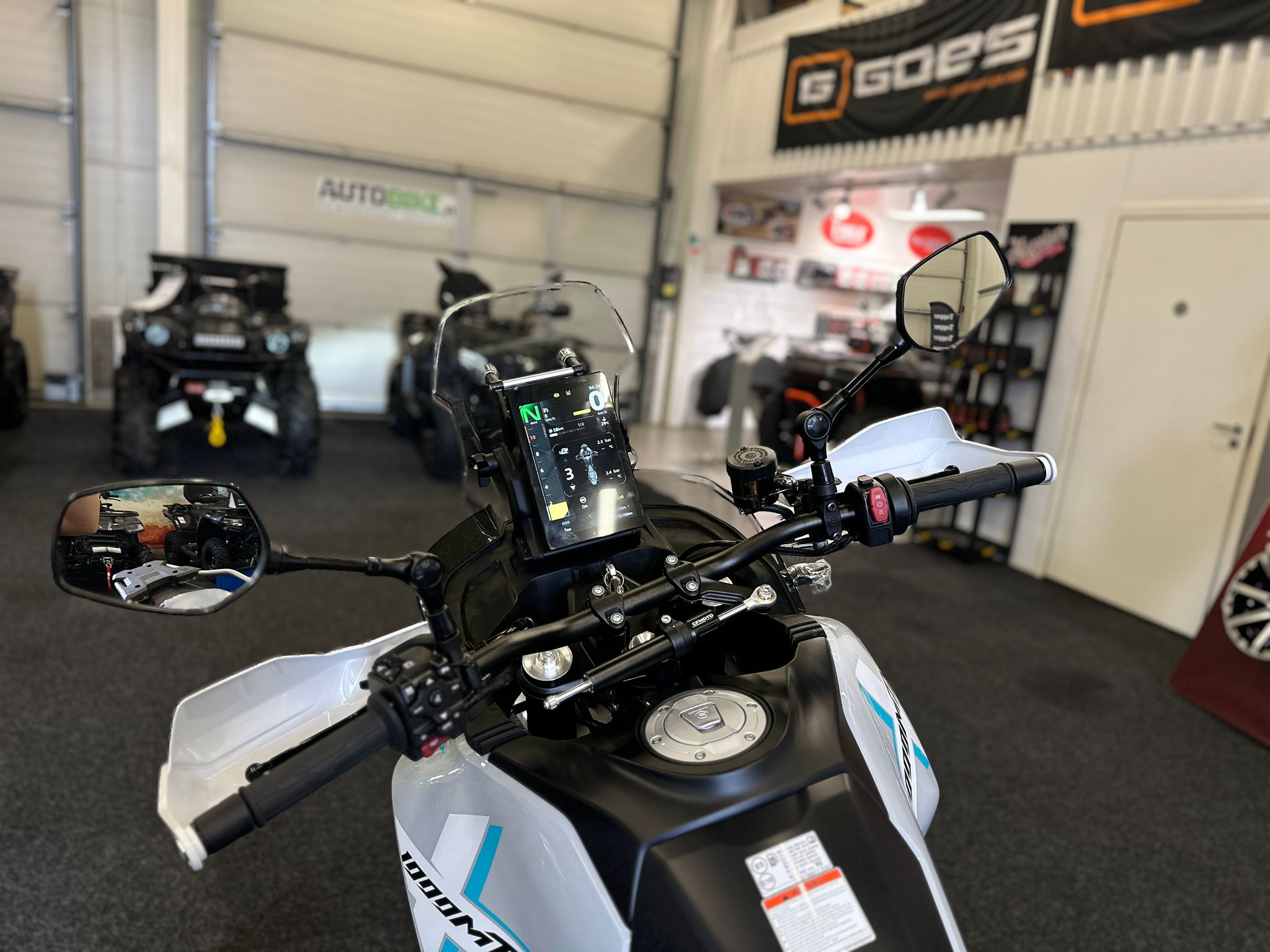 CFMOTO-1000-MT-X-1-18 at 203523 4 - AutoBike Close-up of a motorcycle's handlebars and digital dashboard inside a showroom, with other bikes blurred in the background. - AutoBike