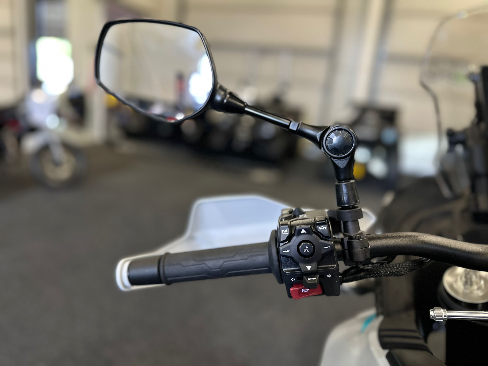 CFMOTO-1000-MT-X-1-18 at 203523 5 - AutoBike Close-up of a motorcycle handlebar with a rear-view mirror and control switches, in a workshop. - AutoBike