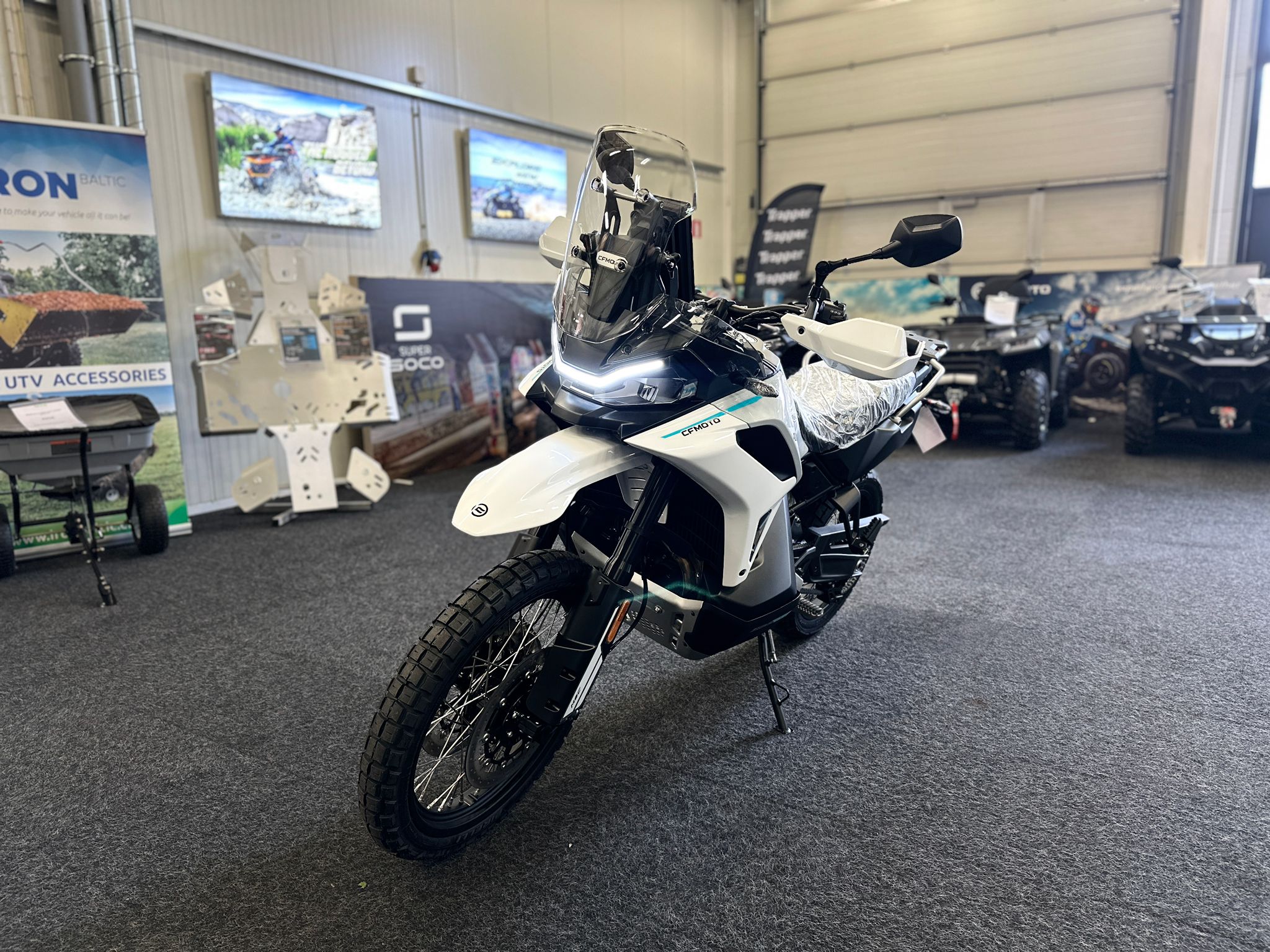 CFMOTO-1000-MT-X-1-18 at 203523 - AutoBike White adventure motorcycle on display in a showroom, with protective windshield and knobby tires visible in the foreground. - AutoBike
