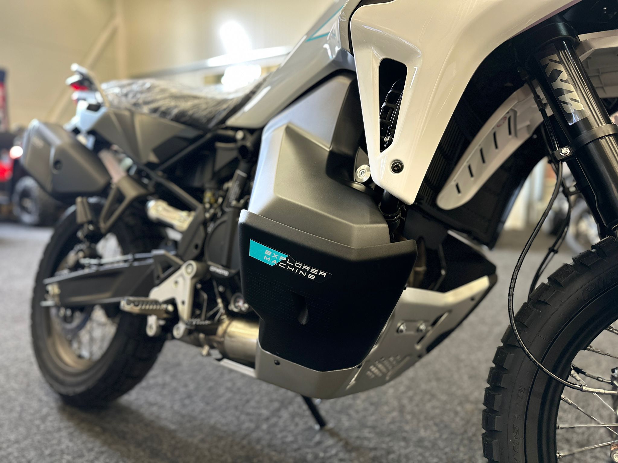 CFMOTO-1000-MT-X-1-18 at 203524 1 - AutoBike Close-up of a white adventure motorcycle's front section, showing the fork, tire, and a turquoise 'Explorer Machine' sticker on the side panel. - AutoBike