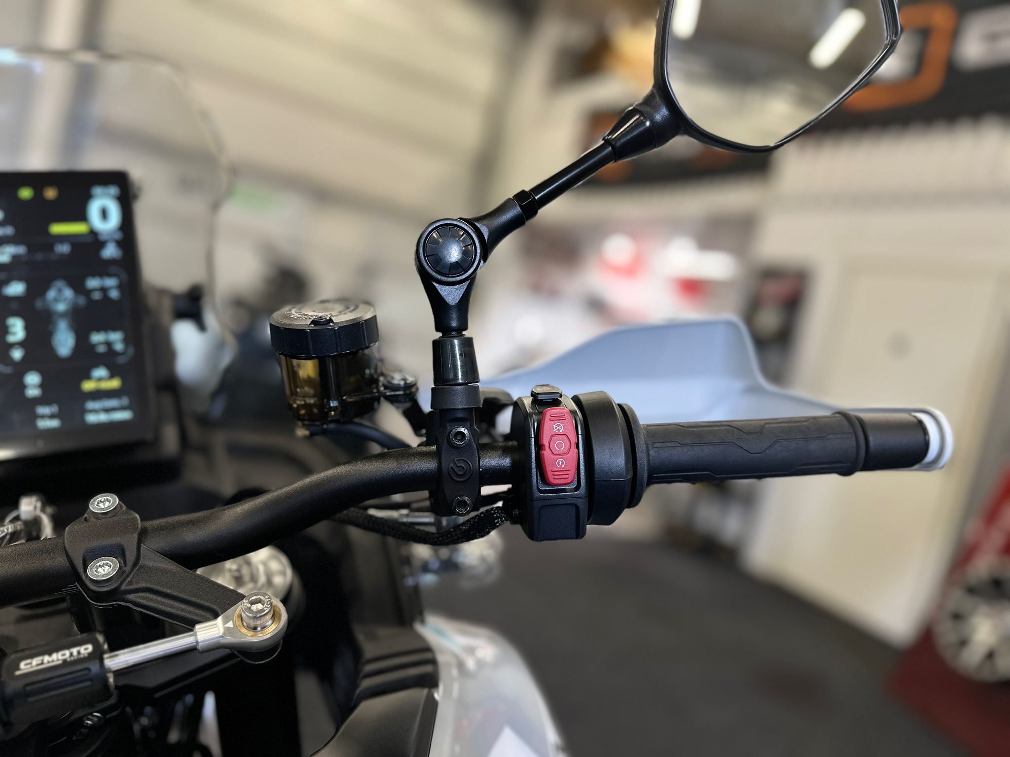 CFMOTO-1000-MT-X-1-18 at 203524 2 - AutoBike Close-up of motorcycle handlebars with a digital dashboard on the left and a rearview mirror above, showing controls and cables in a workshop setting. - AutoBike
