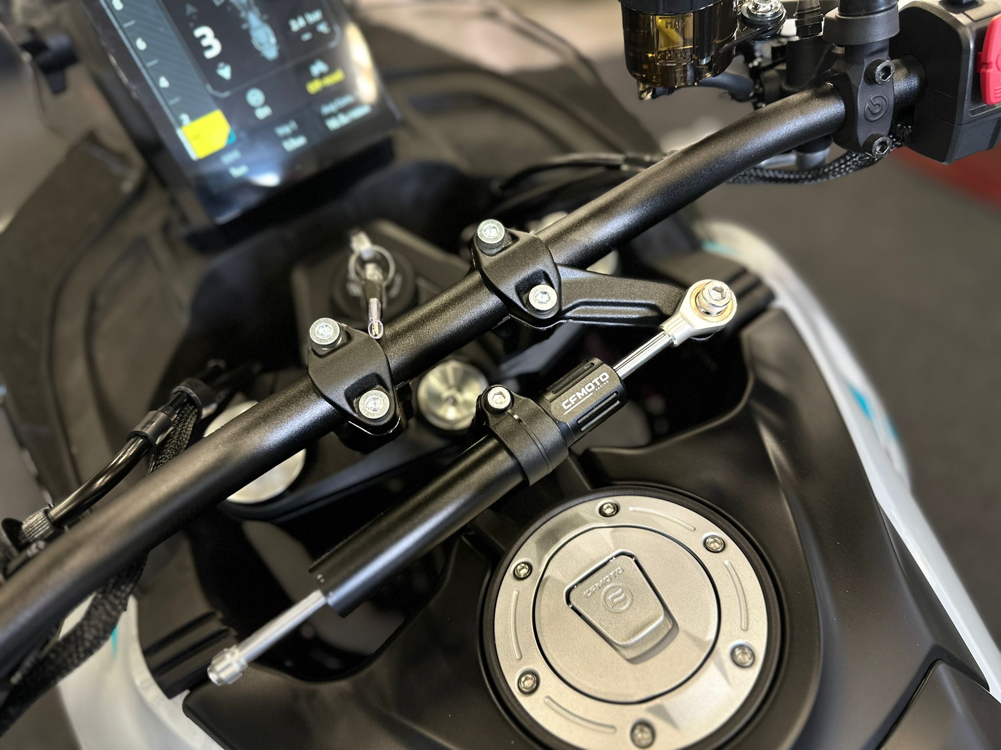 CFMOTO-1000-MT-X-1-18 at 203524 3 - AutoBike Close-up of a motorcycle handlebar area with a digital instrument display and clamp bolts. - AutoBike