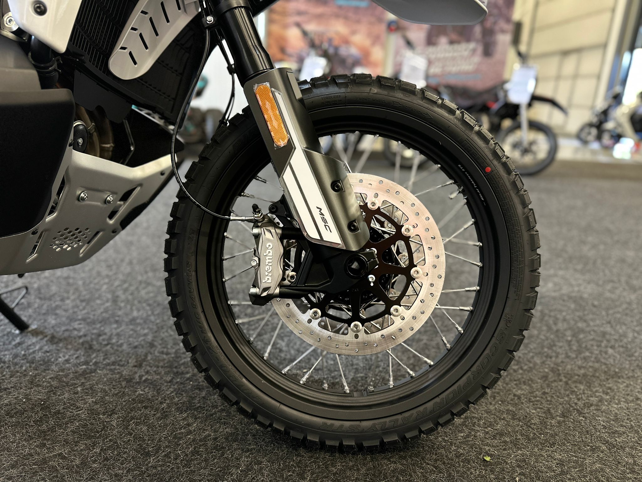 CFMOTO-1000-MT-X-1-18 at 203525 - AutoBike Close-up of a motorcycle's front wheel, disc brake, and treaded tire with an orange fork reflector visible at the fork leg. - AutoBike