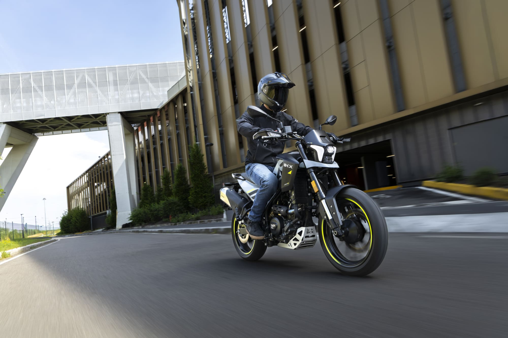 Benelli-bkx-125-s 1 - AutoBike Motorcyclist wearing a helmet and jacket rides a black and yellow motorcycle along a city street beside a modern building. - AutoBike