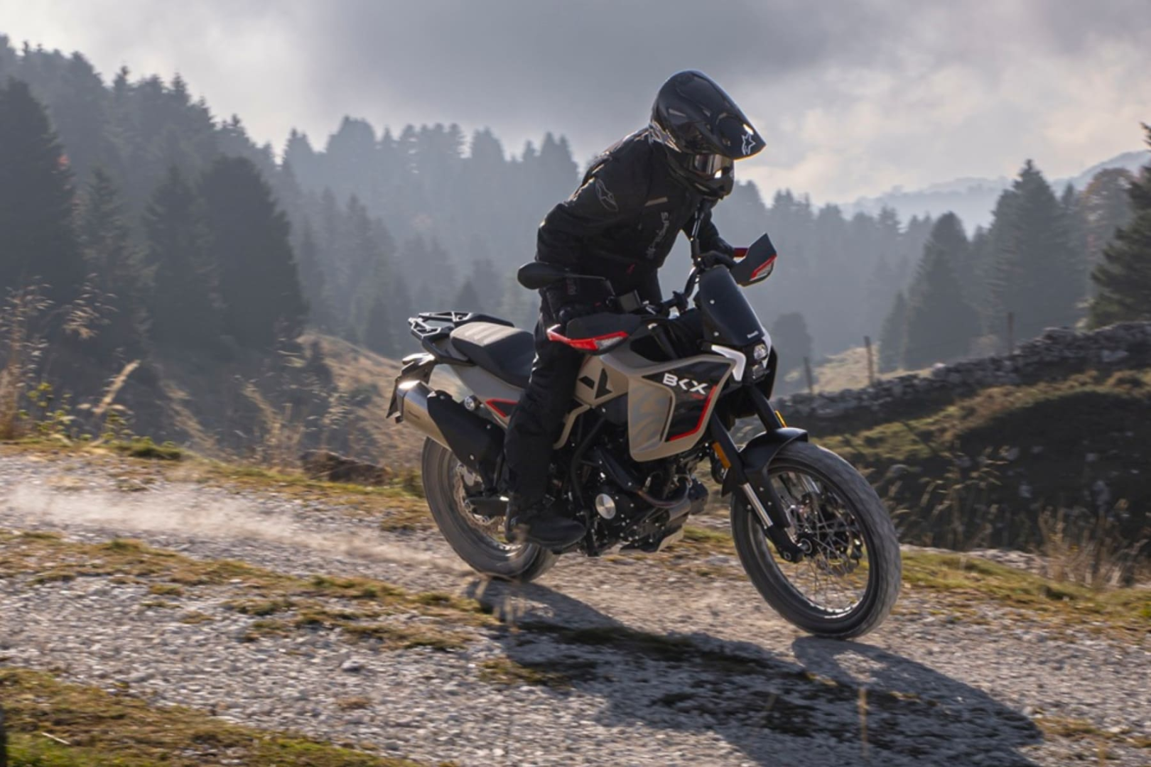 Benelli-bkx-125-s - AutoBike Rider on a dual-sport motorcycle navigating a rocky mountain trail with trees in the background, wearing protective gear and a full-face helmet - AutoBike