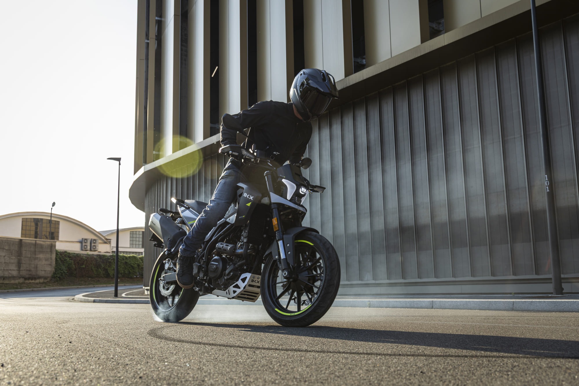 Benelli-bkx-125-s 2 - AutoBike Motorcyclist in black gear performing a wheelie on a sport bike beside a modern glass building, sun flare in the background. - AutoBike