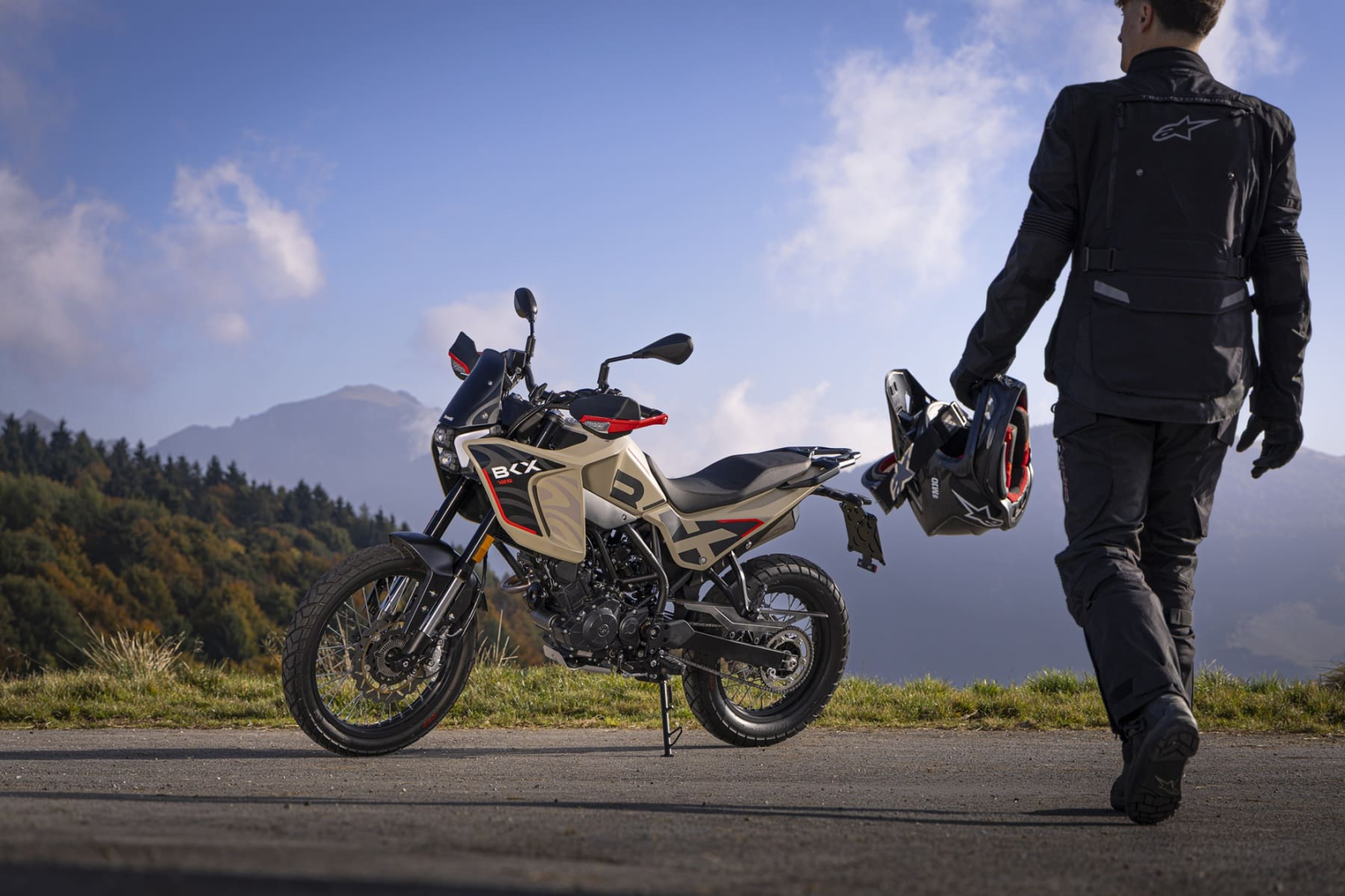 Benelli-bkx-125-s 3 - AutoBike Adventure motorcycle on a hilltop with a rider in protective gear walking toward the bike, helmet in hand, mountain backdrop - AutoBike