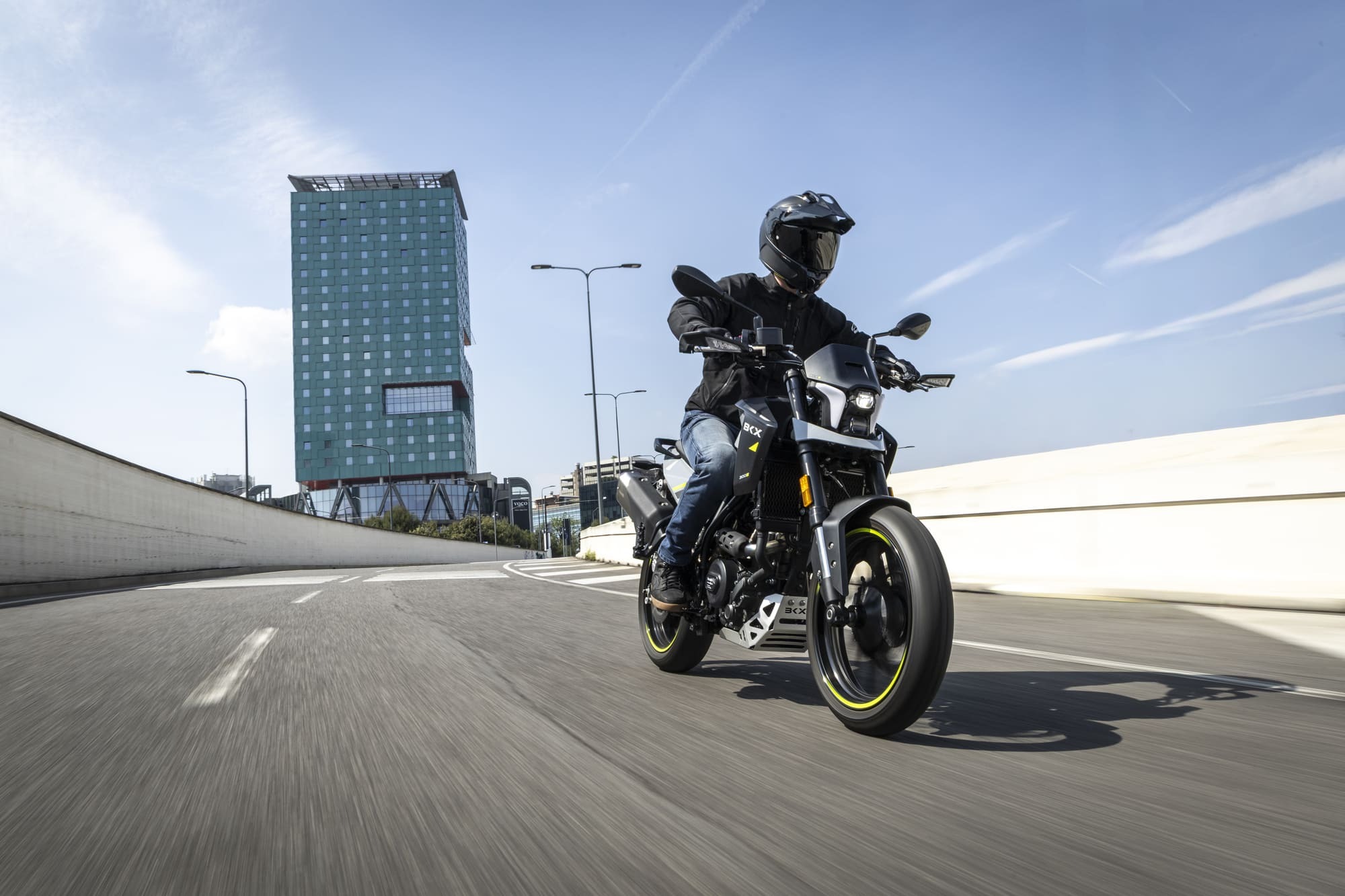 Benelli-bkx-125-s 3 - AutoBike Motorcyclist wearing a helmet and black jacket rides a motorcycle on a curved urban road with a green glass high-rise building in the background. - AutoBike