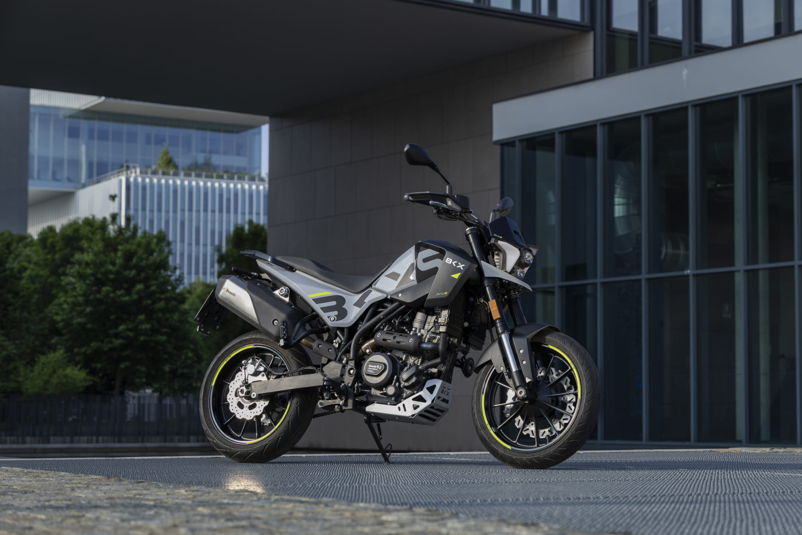 Benelli-bkx-125-s 3 - AutoBike Sport motorcycle parked on a metal surface beside a modern glass building, with greenery in the background. - AutoBike