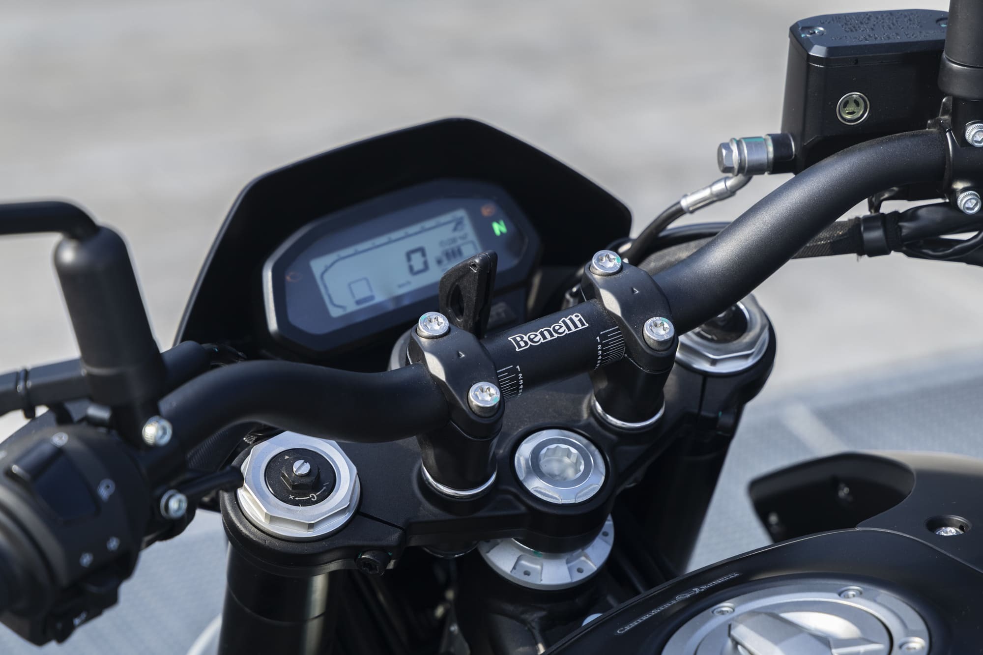 Benelli-bkx-125-s - AutoBike Close-up of a motorcycle cockpit showing handlebars, clamp bolts, and a digital instrument panel with a Benelli logo on the clamp. - AutoBike
