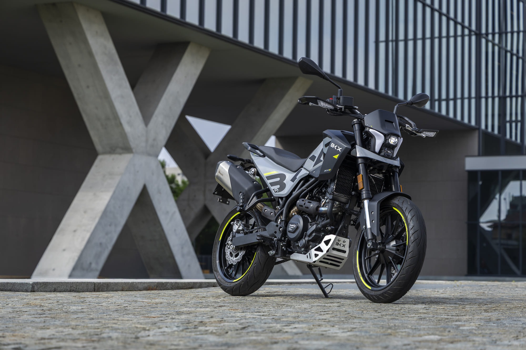 Benelli-bkx-300 4 - AutoBike Sport motorcycle parked on a cobblestone plaza beside a modern building with diagonal concrete supports - AutoBike