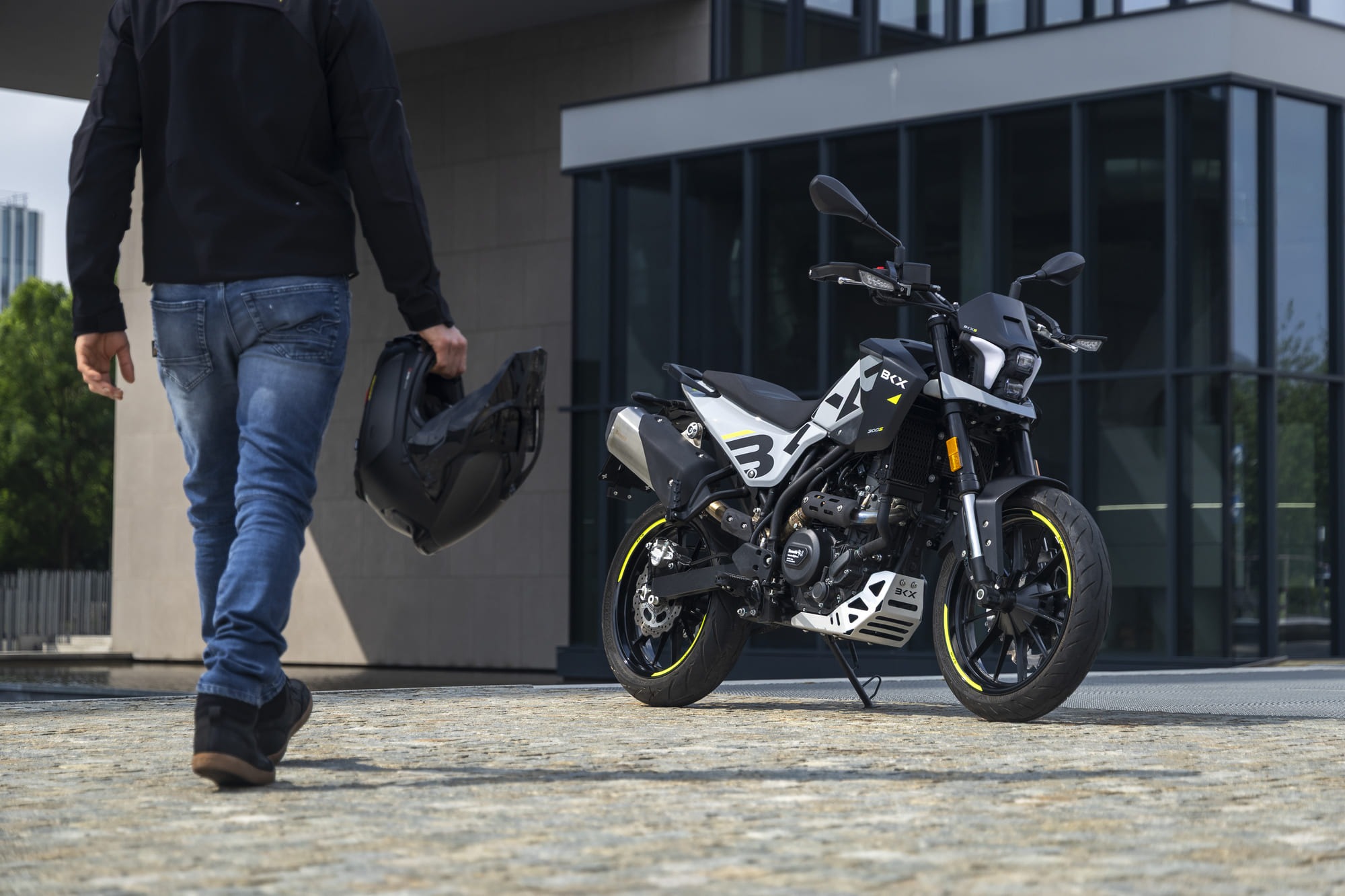 Benelli-bkx-300 6 - AutoBike Person walking away, holding a helmet, beside a white and black motorcycle parked outside a modern glass building. - AutoBike