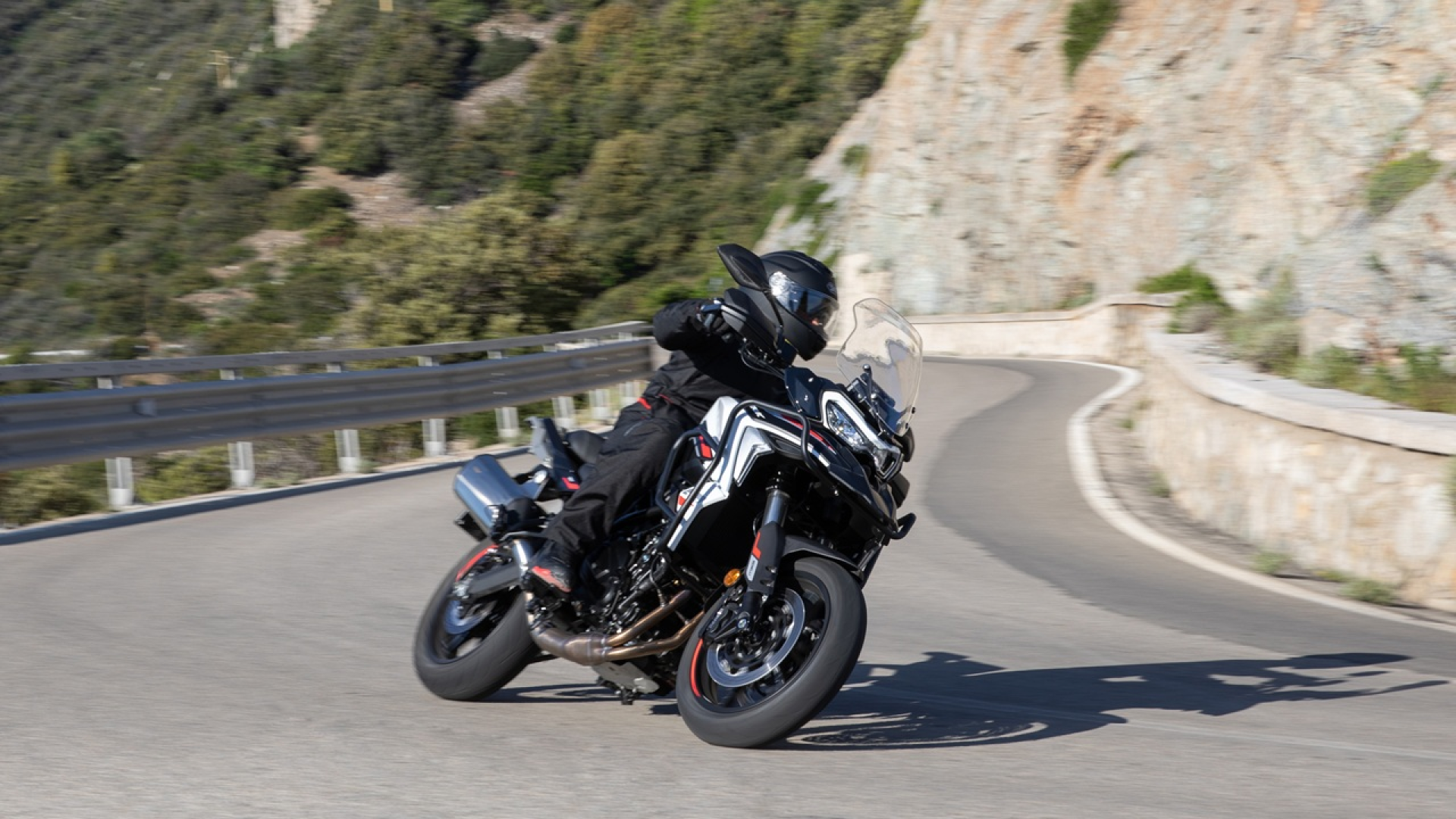 Benelli-trk-702 1 - AutoBike Motorcyclist in black gear leaning into a sharp turn on a curving mountain road with a stone wall on the right and guard rails on the left. - AutoBike
