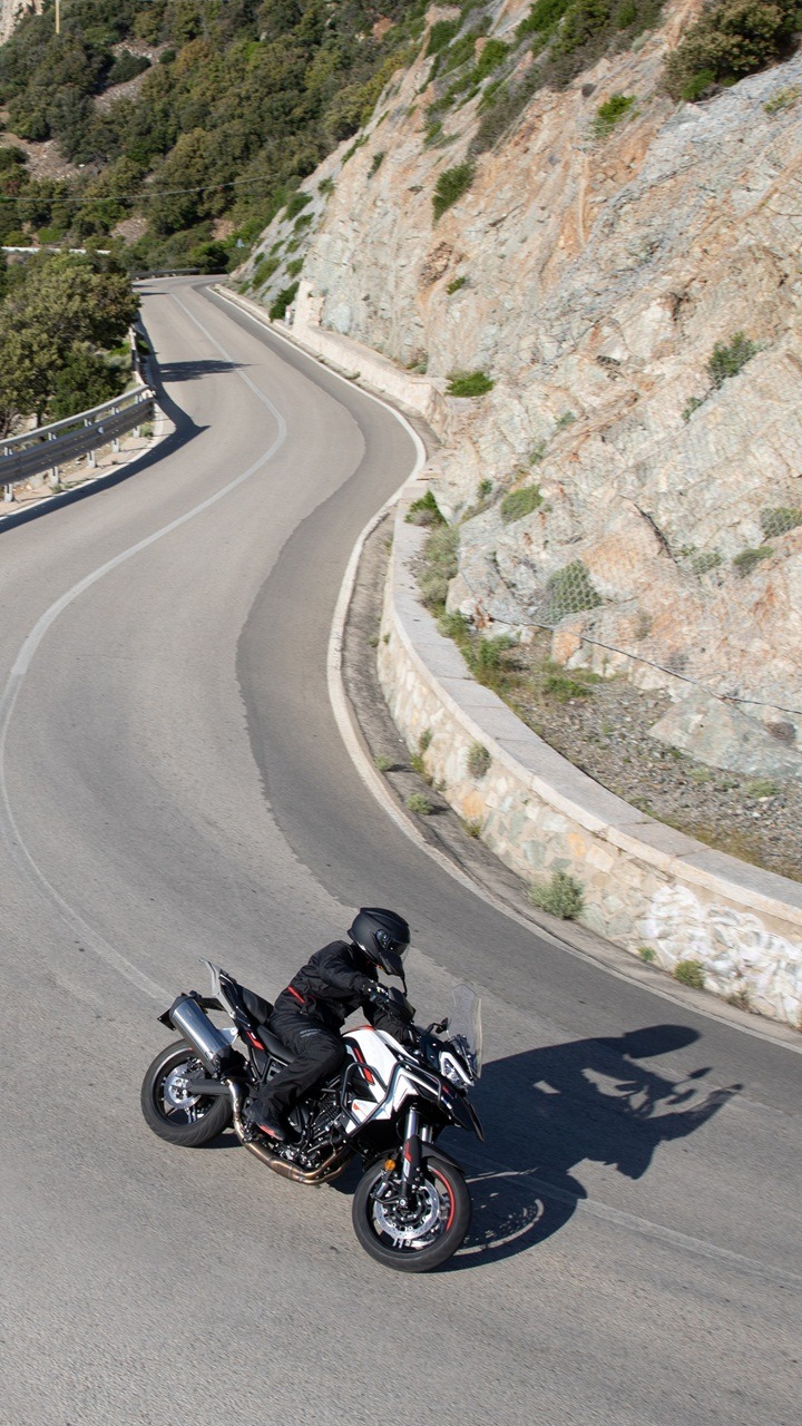 Benelli-trk-702 4 - AutoBike Motorcyclist in black gear on a white sport motorcycle navigating a sharp bend on a mountain road beside a rocky cliff. - AutoBike