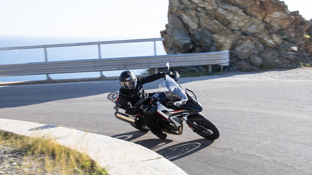 Benelli-trk-702 5 - AutoBike Motorcyclist in black gear leans into a sharp turn on a coastal road, with a metal guardrail and rocky cliff behind and the sea in the distance. - AutoBike