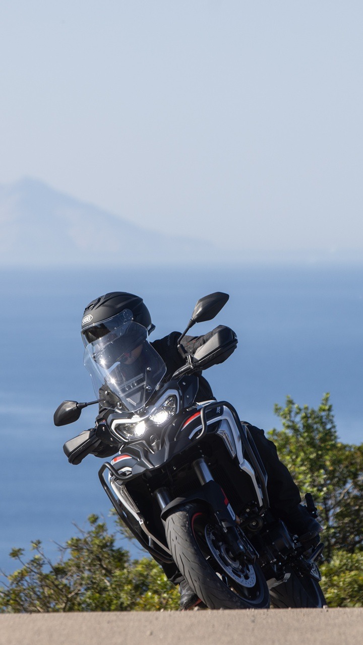 Benelli-trk-702 6 - AutoBike Motorcycle rider in black protective gear leaning into a turn on a coastal road with the sea and hills in the background. - AutoBike