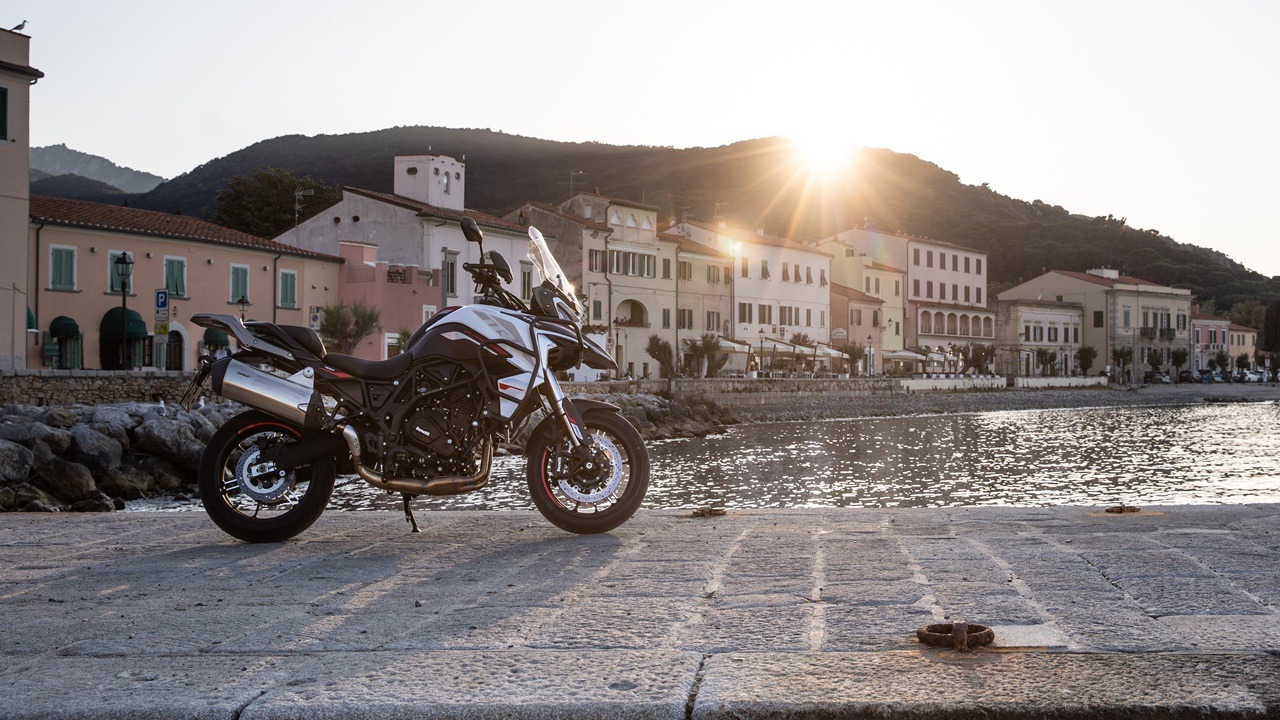 Benelli-trk-702 8 - AutoBike Motorcycle parked on a stone quay by the sea with pastel town buildings and sunlit hills in the background. - AutoBike