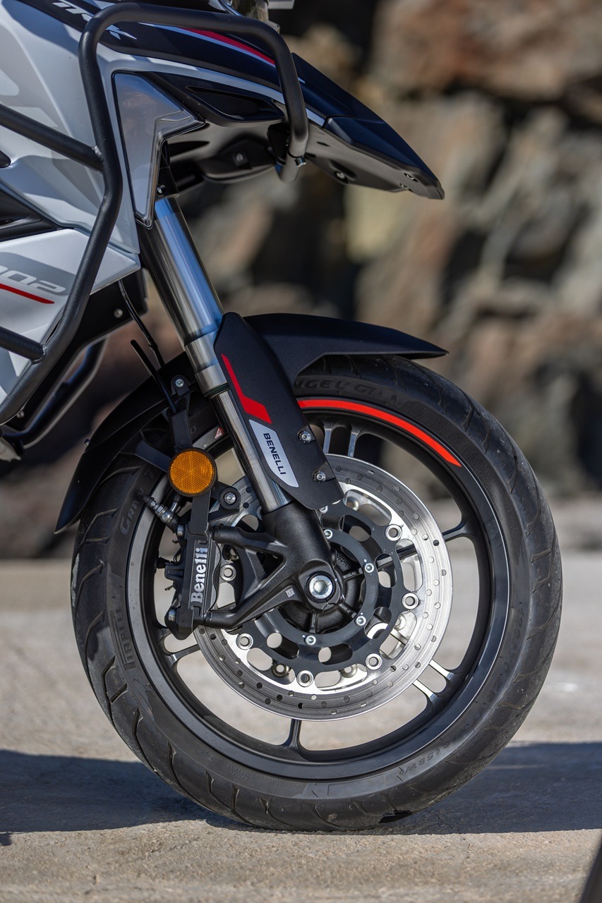 Benelli-trk-702 - AutoBike Close-up of a Benelli motorcycle front wheel with disc brake, black fork, and red-accented fender in a rocky background. - AutoBike
