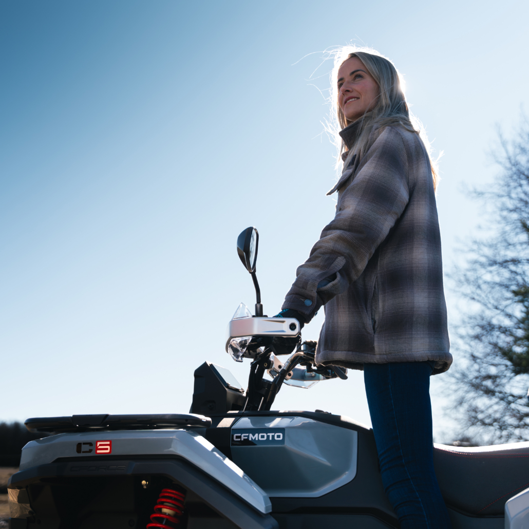CFMOTO_CFORCE-C5_Outdoor_6-1030x1030 - AutoBike Woman in a plaid jacket stands beside a gray CFmoto snowmobile outdoors on a bright sunny day, looking upward. - AutoBike
