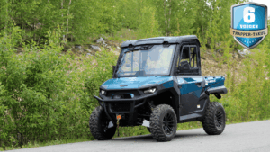 Blue rugged utility terrain vehicle (UTV) on a paved road beside green shrubbery and trees, front-left view, with a large blue badge reading '6 VUODEN TRAPPER-TAKUU' in the top-right corner.