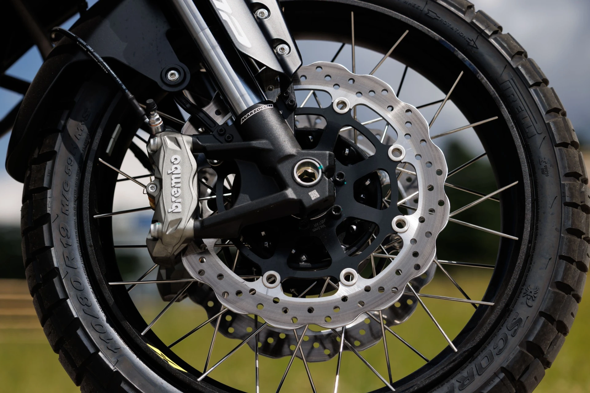 TRK902_Xplorer_041-7 - AutoBike Close-up of a motorcycle front wheel showing a perforated disc brake, Brembo caliper, and spoked rim on an off-road tire. - AutoBike