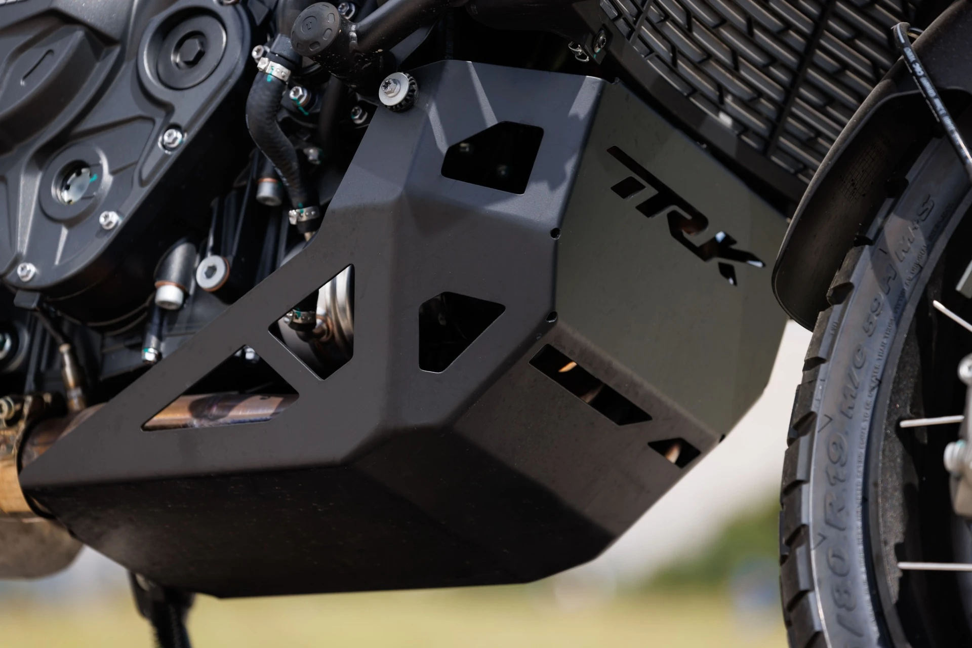 TRK902_Xplorer_049-0 - AutoBike Close-up of a motorcycle's lower frame and front wheel, showing metal skid plate and engine components - AutoBike