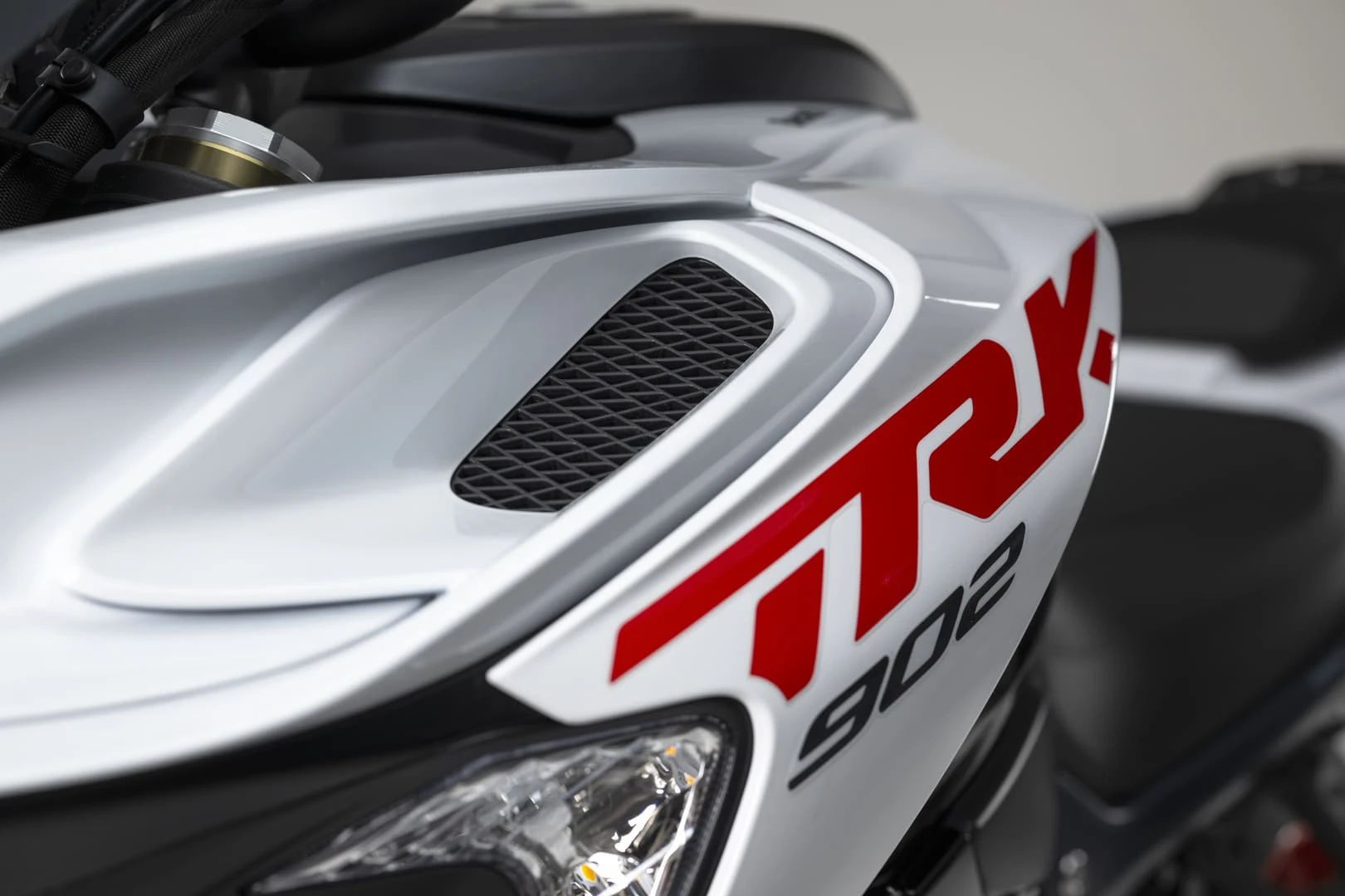 trk902stradale_12 - AutoBike Close-up of a white sport motorcycle front fairing with a black mesh vent and bold red graphics, partial headlight in the lower left corner. - AutoBike