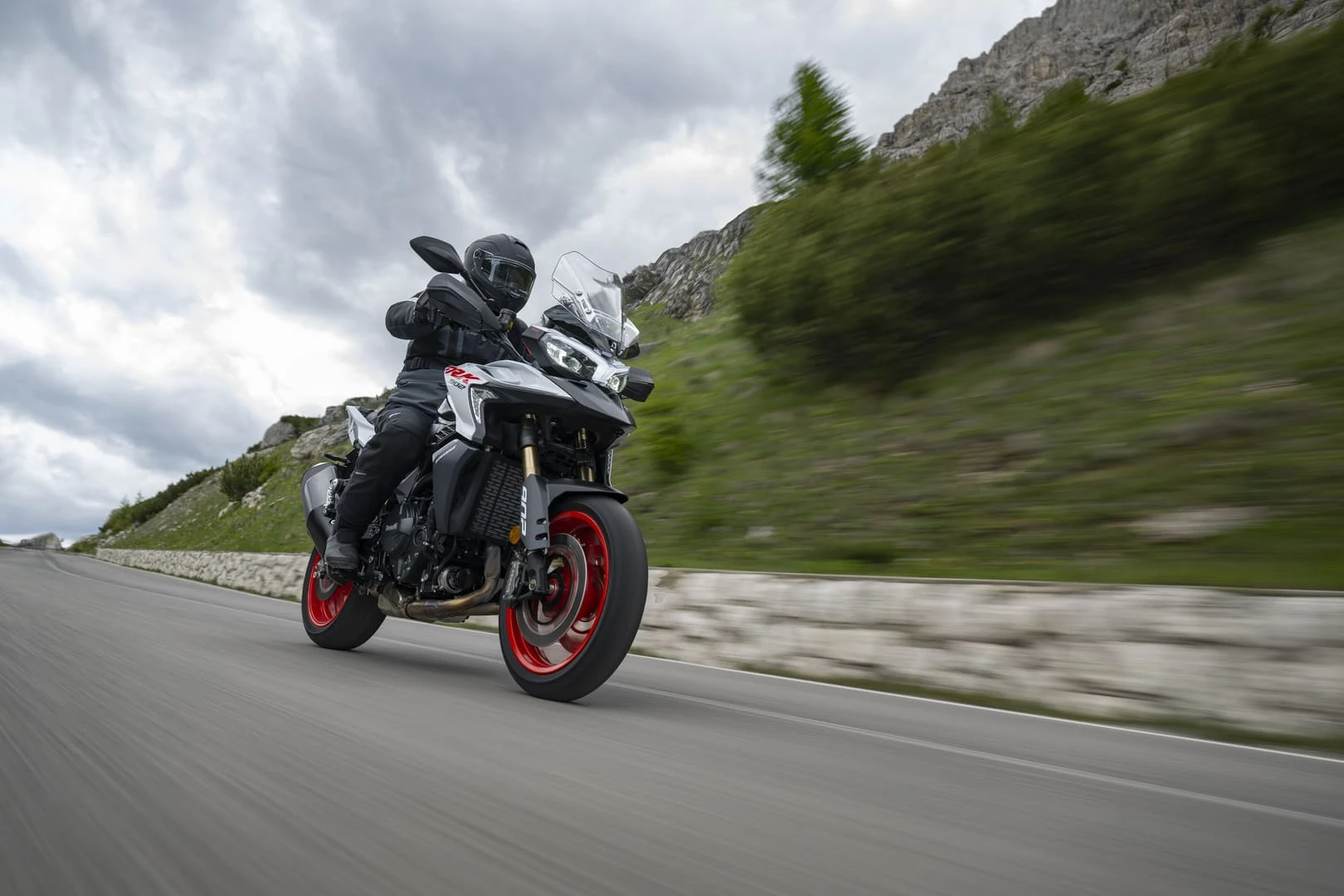 trk902stradale_action_02 - AutoBike Motorcyclist wearing black gear rides a rugged adventure bike on a windy mountain road with a stone barrier and rocky hillside ahead - AutoBike