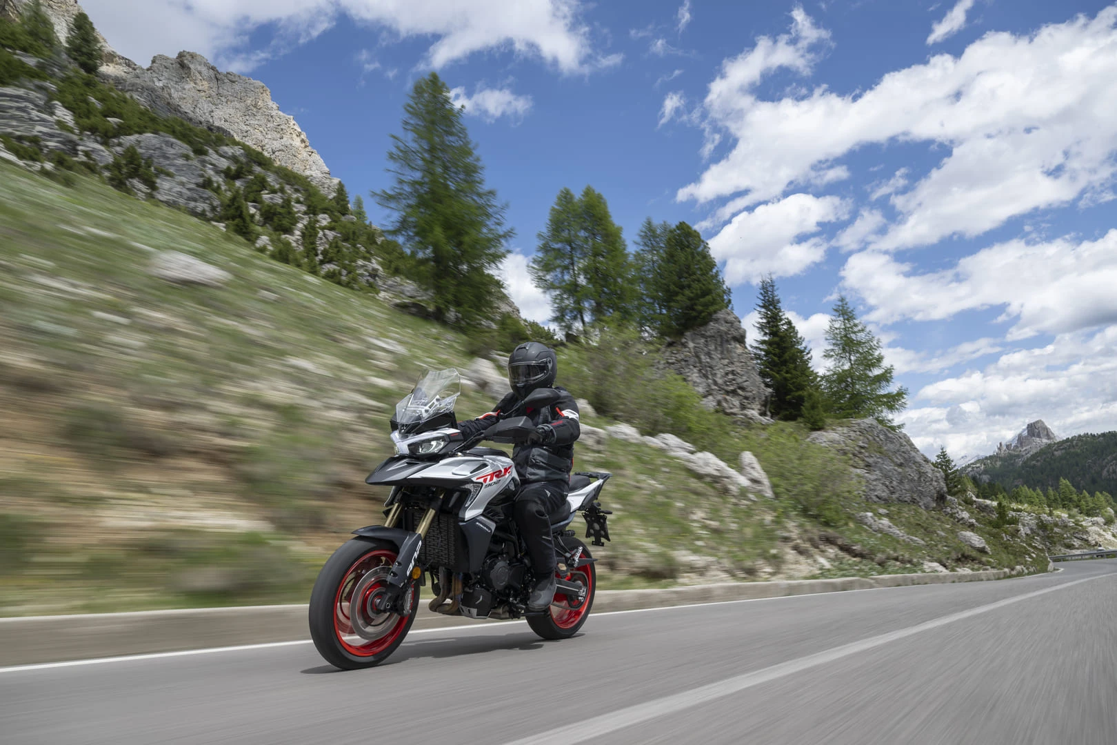 trk902stradale_action_06 - AutoBike Motorcyclist in black protective gear riding a silver adventure motorcycle on a winding mountain road, with rocky cliffs, pine trees, and a blue sky in the background. - AutoBike