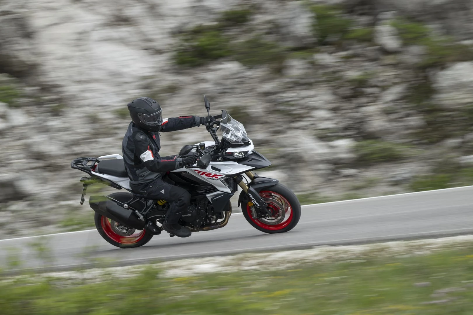 trk902stradale_action_10 - AutoBike Motorcyclist in full protective gear leaning through a curve on a white-and-black sport bike with red wheels on a winding mountain road backdrop. - AutoBike