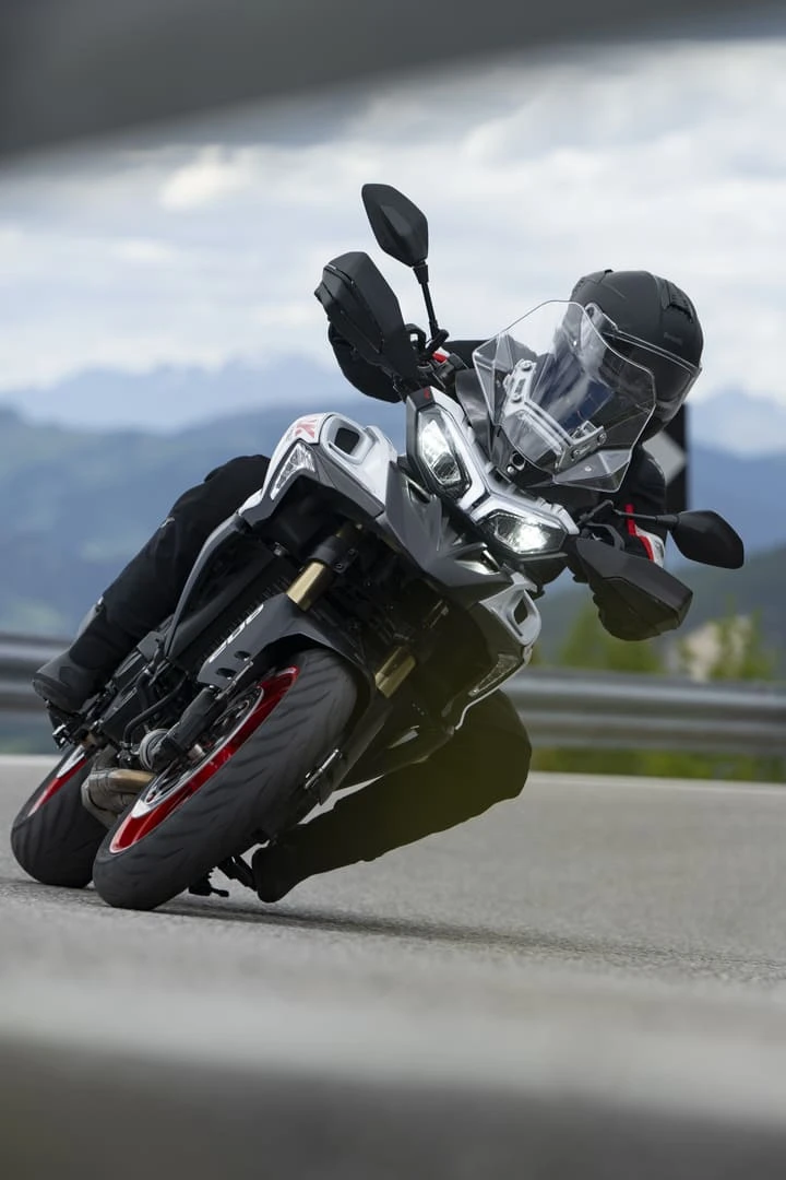 trk902stradale_action_12 - AutoBike Motorcyclist leaning into a turn on a sport-touring bike with a clear windshield and protective gear, mountainous backdrop. - AutoBike