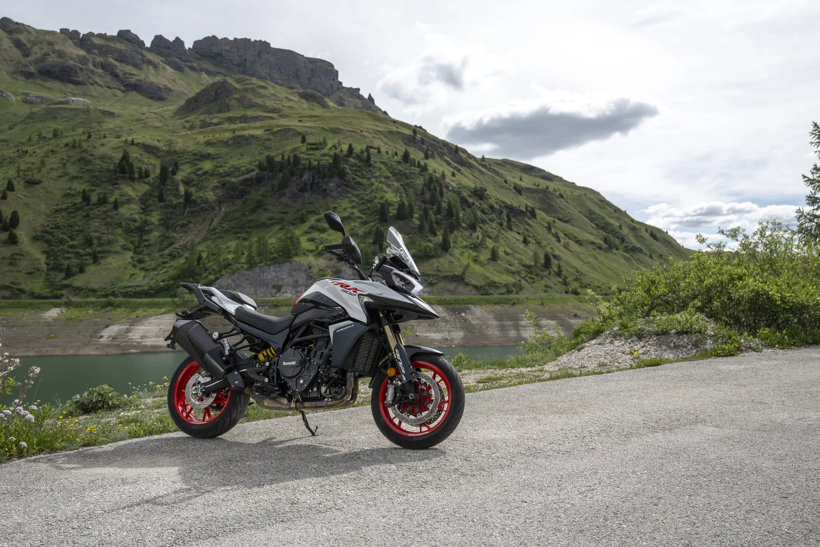 trk902stradale_ambient_01 - AutoBike Sport motorcycle with red rims parked on a roadside, green hills and rocky mountains in the background under a cloudy sky. 125 chars? (concise alt) - AutoBike