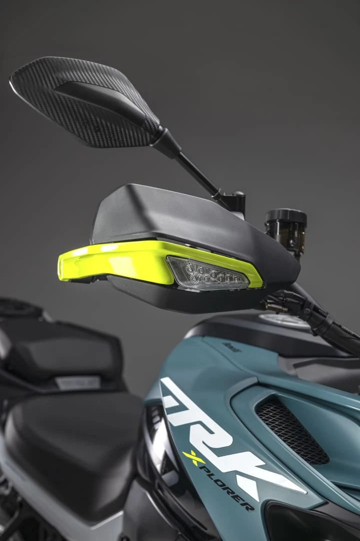 trk902xplorer_29 - AutoBike Close-up of a motorcycle handlebar with a carbon-fiber rearview mirror, neon yellow front fairing, and teal body panel with 'Xplorer' branding visible on the side. - AutoBike