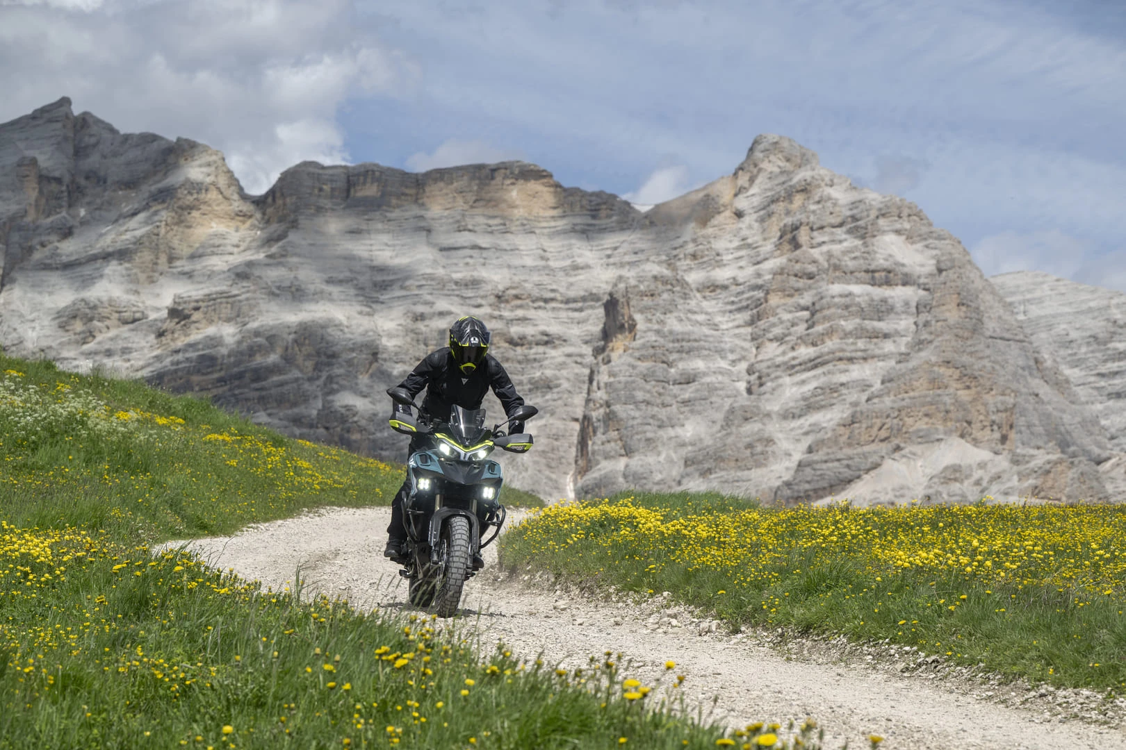 trk902xplorer_action_01 - AutoBike Rider in black gear on a dirt bike along a gravel path through a field of yellow flowers, with rocky mountains in the background. - AutoBike