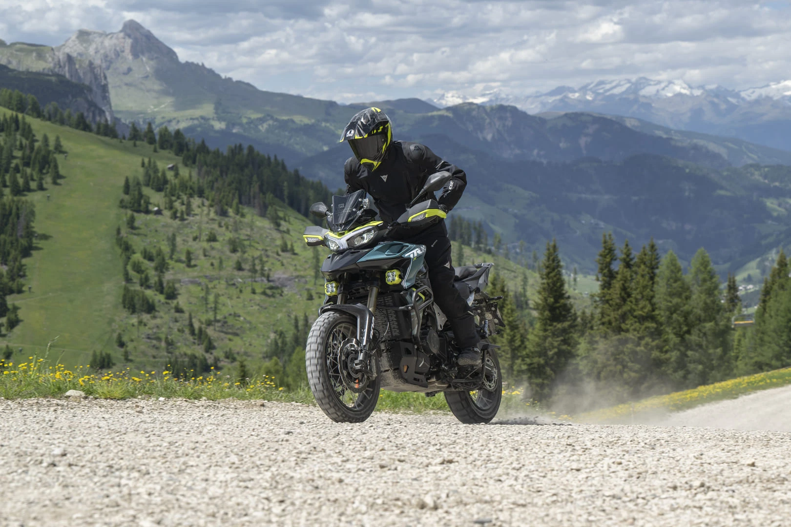 trk902xplorer_action_03 - AutoBike Motorcyclist in black protective gear with a neon-yellow helmet riding a green adventure motorcycle on a gravel mountain road with pine trees and distant snow-capped peaks in the background. - AutoBike