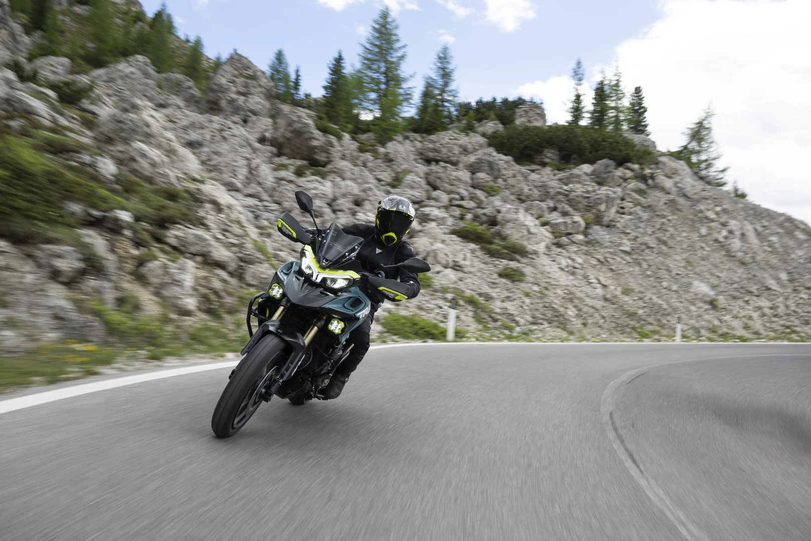 trk902xplorer_action_10 - AutoBike Motorcyclist wearing black gear and helmet leans into a sharp turn on a winding mountain road with rocky hillside and pine trees in the background, neon-yellow accents visible on the bike and gear. - AutoBike